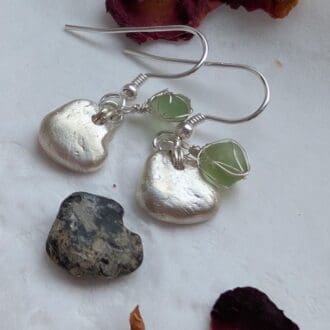 A pair of dainty silver heart earrings each with a tiny piece of pale green sea glass wrapped in very fine silver wire