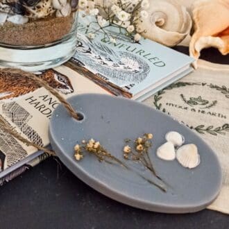 Coast grey soy wax melt and wardrobe freshener with small white shells and dried white flower embeds sits on a book next to the Coast botanical candle surrounded by shells.