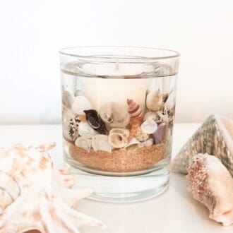 Coast botanical candle with natural coloured sand and shells rests on a surface between two large seashells