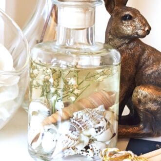 Coast luxury botanical diffuser. Clear fresh scented fragrance oil in a 200ml apothecary bottle with shells and small white dried flowers suspended in the liquid. The reed diffuser is placed on a white surface with a large white flower and a bronze hare ornament.