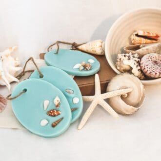Set of 3 Blue Sea oval shaped soy wax melts/ room fresheners in an aqua marine colour embeded with small shells and a twine loop for hanging. The sea inspired wax melts are on a white background resting on a card gift box surrounded by large shells, a starfish and a neutral coloured bowl spilling more shells.