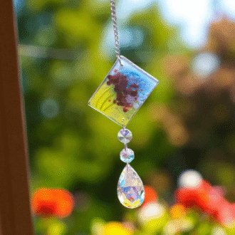 Handmade fused glass lavender suncatcher with teardrop crystal, hanging in a sunny window with a blurred flower garden in the background.