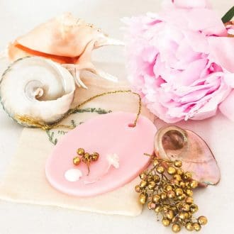 Pink oval shaped soy wax tablet with gold hanging loop, decorated with gold, white and pink botanicals. The wax melt/room freshener is on a white background resting on a muslin bag, surrounded by a large pink peony, gold berries, mother of pearl and large sea shells which give an air of relaxation and luxury.
