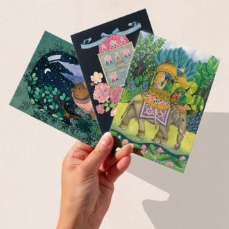 A hand holding three hand-illustrated art greeting cards featuring The Illustrarium’s original designs: a bear under a starry sky in a glass terrarium, a dolls’ house surrounded by peonies, and an Indian elephant carrying a girl and a monkey through tropical greenery.