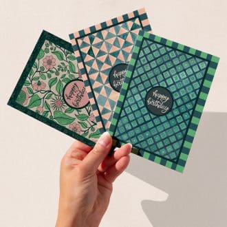 A hand holding three hand-illustrated birthday cards featuring The Illustrarium’s original designs: Regency-inspired floral, green geometric squares, and pink triangles — each with “Happy Birthday” in the centre.