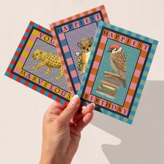 A hand holding three colourful greeting cards featuring The Illustrarium’s original animal designs: a leopard with “You’re Marvellous,” a crowned chihuahua with “Happiest Birthday,” and a sparrow perched on books with “Happiest Birthday.”