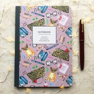 A hand-painted A5 notebook featuring chihuahuas and vintage-style fashion accessories on a pink background, styled with a red fountain pen and delicate fairy lights.