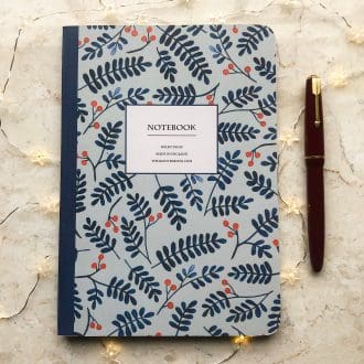 A blue A5 notebook featuring hand-painted navy leaves and red berries, styled with a red fountain pen and vintage papers on a wooden desk.