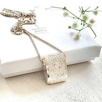 A fine silver pendant, square design with a textured pattern and a white cz stone in the centre. The pendant has a sterling silver chain and is displayed on a white background leaning on a white gift box.