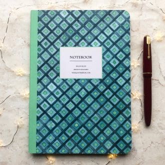 A hand-painted geometric A5 notebook with a mint green spine and a blue and green diamond pattern, styled with a red fountain pen and fairy lights on a marble surface.