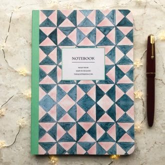 A hand-painted geometric A5 notebook featuring pink and teal triangles with a mint green spine, styled with fairy lights and a red fountain pen on a marble background.