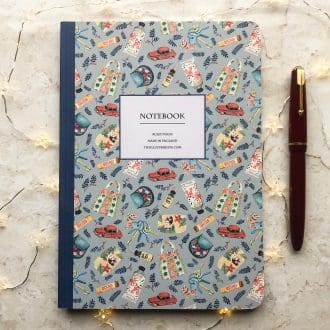 A beautifully illustrated A5 notebook featuring hand-painted fashion and vintage-inspired designs, including handbags, perfume, and flowers, shown with a red pen and warm fairy lights on a marble background.