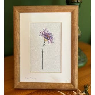 A brown frame standing on a table featuring a single stem purple embroidered cornflower