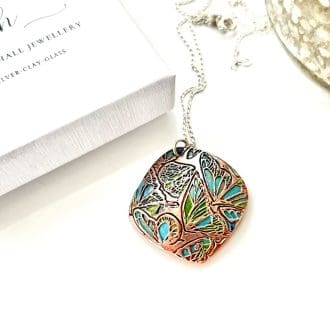 A handcrafted copper pendant in a soft diamond shape and a textured pattern that has been hand-painted in turquoise and green patina paints. The pendant has a sterling silver chain and is displayed on a white surface.
