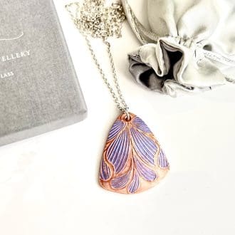 A handcrafted copper pendant which is textured with a flower pattern hand painted in lilac metallic paints. It has a sterling silver chain and is displayed on a white background.