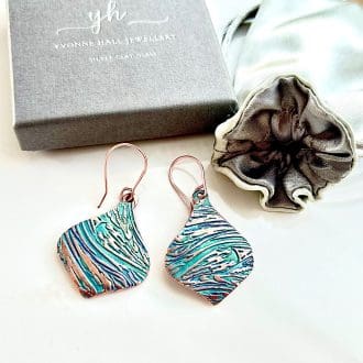 A pair of handcrafted copper drop earrings in a round shape with a swirl textured pattern. They are hand-painted in soft aqua and pale turquoise and have copper ear sires, displayed on a white background with a grey gift box and satin jewellery pouch above them.