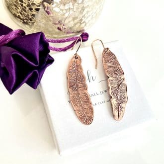 A pair of copper drop earrings in a long textured tribal feather design with a lot of intricate detail and with copper ear wires. They are displayed on top of a white gift box.