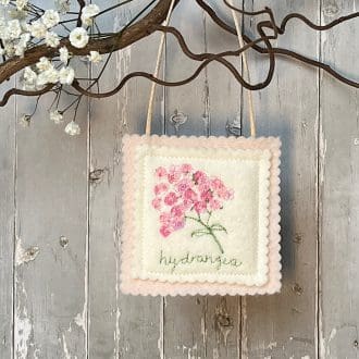 Small square felt decoration hangs from a branch. Pink and cream it features an embroidered pink hydrangea. The word hydrangea is embroidered underneath the flower