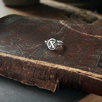 A silver ring with the letter X lies on a book with an old brownish red cover.