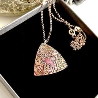 A triangular shaped copper pendant with a textured pattern that has been hand-painted in greens and burgundy metallic paints, it has a rose gold plated chain. It is displayed resting on black velvet inside a grey gift box.