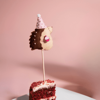 Handmade felt hedgehog cake topper, hedgehog is wearing a pink party hat with a pom pom and is carrrying a cupcake ready for a birthday party.