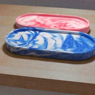 Two oval trinket trays next to each other on a light wooden countertop. The tray nearest to the front is blue marble, the one next to it is red marble.