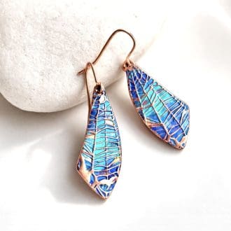 A pair of handcrafted copper drop earrings with a spider web design hand-paonted in metallic blues and turquoise paints. They are in a wing shape and are displayed resting on a white stone with a white background.