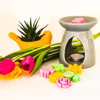 Handcrafted soy wax melts with a floral fragrance in pink, green and yellow hues rest in front of a wax burner next to tulips and daffodils with a bright yellow bird planter in the background.