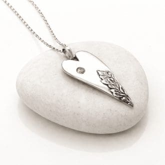 A handcrafted silver elongated heart necklace with a leaf texture at the bottom and a amber colourd CZ near the top. The necklace has a sterling silver bail and chain and is displayed resting on a light colured smooth stone.