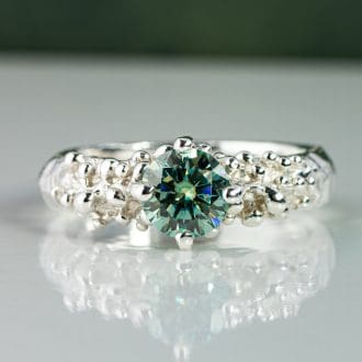 Handmade sterling silver ring with a textured organic band, set with a round green moissanite gemstone, photographed in soft natural light with a subtle reflection.