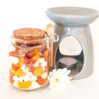 Glass jar of soy wax scoopies in white, yellow and orange with a cork lid and wooden scoop stands on a white background next to a grey wax burner and white flower.