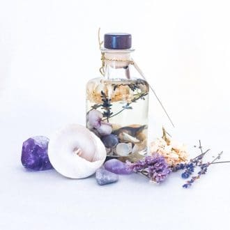 Luxury lavender reed diffuser with shells and dried flowers in an apothecary style bottle, on a white surface surrounded by purple and white flowers, a large white shell and purple semi-precious stones.