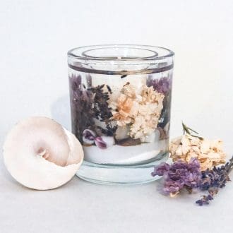 Lavender soy and gel wax refillable botanical candle with flower and shell embeds on a white surface next to a large shell and dried flowers in white and purple.