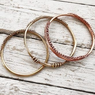 Three handcrafted copper and bronze bangles with a unique textured pattern around the outer edge. The bangles are displayed on a white background resting on a light coloured stone.