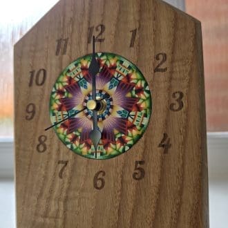 Handcrafted solid oak mantel clock with laser-engraved numerals and colourful millefiori-inspired polymer clay centrepiece artwork by Bridget Derc, featuring black hands and sharp chamfered edges.