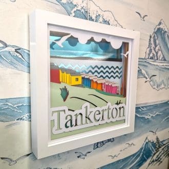 A layered photo artwork of Tankerton slopes with the beach huts overlooking the sea. It has a mirrored blue background and the word Tankerton written at the bottom of the front layer. It is seen from the left edge and is in a white box frame