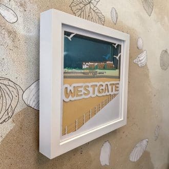 A layered paper artwork of Westgate beach in Kent. It looks from the promenade across the beach towards the Café and homes on the cliff. It is seen from the left edge and is in a white box frame