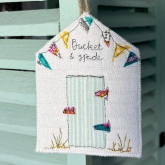 Hanging decoration in the shape of a beach hut with colourful bunting and the words bucket and spade above the door. Hangs on a blue louvre door.